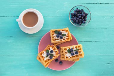 Krem peynirli waffle ve pembe tabakta yaban mersini, turkuaz ahşap masada kahve fincanı. Üst manzara, düz uzanma.
