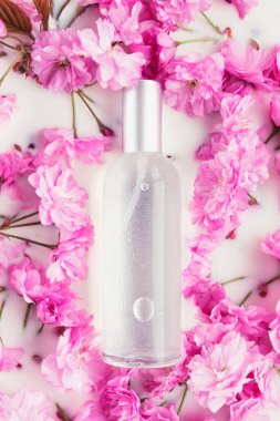 Beyaz suda veya sütte pembe bahar çiçeklerinin arka planında cam bir şişe kozmetik tonik. Doğal organik kozmetik kavramı. Üst görünüm, düz görünüm, maket.