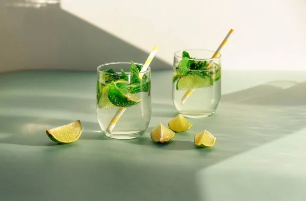 Two glasses of lemonade with straw and lime slices on blue table. Cold refreshing drinks against white wall in sunlight — Stock Photo, Image