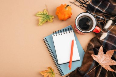 Kahve fincanı, not defterleri, kalem, balkabağı, yapraklar ve bej arka planda ekose eşarpla sonbahar yatıyordu. Sıcak mevsimlik çalışma alanı konsepti. Üst görünüm, model.