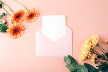 Envelope with blank card, orange gerbera flowers and yellow chrysanthemums on coral pink background. Floral mockup for message or invitation. Top view, flat lay.