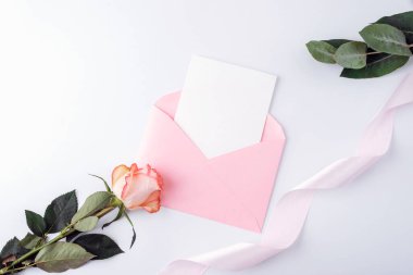 Üzerinde boş kart olan pembe bir zarf, beyaz arka planda gül çiçeği ve kurdele. Tatil tebrik kartı, davetiye, düğün konsepti. Üst görünüm, düz görünüm, model.