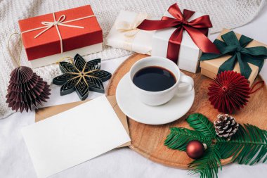 Tahta tepside bir fincan sade kahve ve hediye paketleri, kurdeleler, süsler, kozalaklar ve sonsuz yeşil dallarla çevrili boş bir kart doğal renklerde sıcak bir bayram ortamı yaratıyor. Üst görünüm, model.
