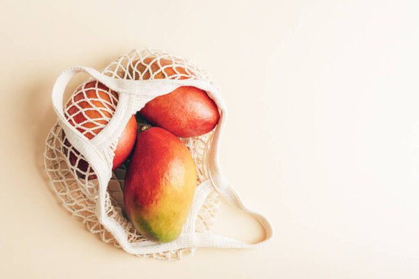 Fresh tropical mango fruits in a cotton mesh bag on a light warm background. Top view, flat lay, copy space.