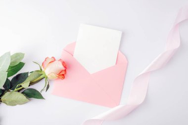 Üzerinde boş kart olan pembe bir zarf, beyaz arka planda gül çiçeği ve kurdele. Tatil tebrik kartı, davetiye. Üst görünüm, düz görünüm, model.