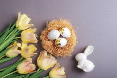 Süslü yuvada paskalya yumurtaları, sarı laleler ve pastel arka planda beyaz seramik tavşanlar. Üst manzara, düz uzanma.