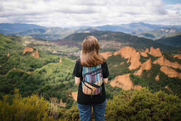 Unrecognizable woman with colorful backpack resting on a tree looking at the forest