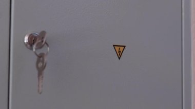 Uyarı sembollü bir elektrik dolabının yakından görüntüsü, yavaş yavaş anahtar deliğine yaklaşıyor, endüstriyel ortamlarda elektrik şoku riskini vurguluyor