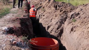 Turuncu yelek giyen işçiler su sistemi genişlemesi için aktif bir çukur kazarken, iş arkadaşları arka planda gözlem ve yardım yaparak işbirlikçi çabaları vurguluyor