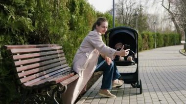 Açık renk ceketli bir kadın, bir bebek arabasının yanındaki bankta otururken akıllı telefonuyla bir anı yakalamayı seviyor. Bağlantı ve neşeyi vurguluyor. Kamera hareketi takip ediyor.