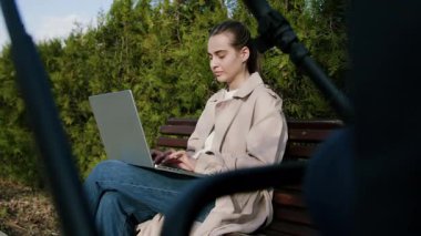 Trençkot giymiş bir kadın, yemyeşil yemyeşil bir bankta otururken aktif olarak dizüstü bilgisayardan online alışveriş yapıyor ve sakin bir doğa deneyimi sergiliyor.