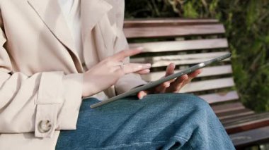 Bej ceketli kadın kullanıcı, parktaki bir bankta tabletle meşgul, çevrimiçi alışveriş eylemlerini vurguluyor, el hareketlerinde kameralar yakınlaşıyor ve cihaz etkileşimi