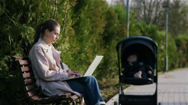 Uzaktan kumandalı bir dizüstü bilgisayarda çalışan kadın, yakınlardaki bir bankta bebek arabası, doğanın ve verimliliğin bir karışımını vurguluyor, kamera çekim sahnesini yakalamak için tavalar