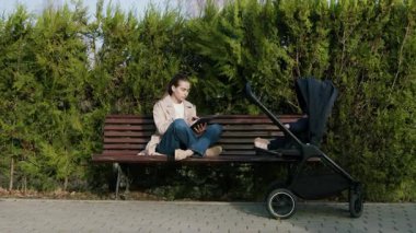 Bir bankta huzurlu bir anın tadını çıkaran bir kadın, yanında bebek arabası olan bir kitap okuyor, yemyeşil bir arazi ile çevrili, sakin atmosferi ortaya çıkarmak için kamera tavaları.