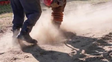 İşçi bir toprak sıkıştırıcı kullanıyor, yol onarımı için toprağı sıkıştırıyor, toz içinde dönüyor, inşaat çalışmalarının ve iş makinelerinin yoğunluğunu vurguluyor