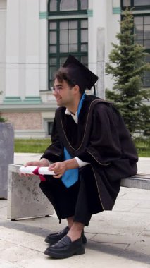 Mezuniyet kıyafetleri, siyah önlüğü ve şapkası içinde üniversite kampüsünün avlusunda bir sandalyede oturan ve mezuniyet diplomasını gururla elinde tutan mutlu bir mezun. Uluslararası çalışmalar