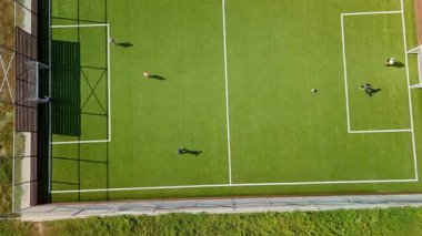 Çocuklar parlak yeşil bir sahada futbol oynuyorlar, takım çalışmasının ve hareketinin heyecanını yakalıyorlar ve topa vurarak kaleye doğru koşuyorlar.