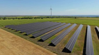 Yeşillik bir alanda sıralanmış güneş panellerinin hava görüntüsü, yenilenebilir enerji teknolojilerinin doğal bir manzaraya entegrasyonunu gösteren bir rüzgar türbini tarafından tamamlanmış.