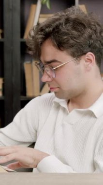 Portrait of a student boy browsing and reading a book during a break between lessons. Student with glasses reads a book in the modern library of the university campus.Modern conditions for students