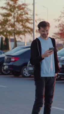 Casual-dressed man is standing in a shopping district parking lot, using his smartphone, with parked vehicles and colorful trees creating a warm evening ambiance
