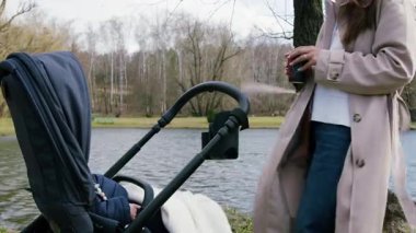 Bej ceketli bir kadın bebek arabasının yanında duruyor, elinde bir içecek var, kamera sakin park atmosferini yakalamak için tava gibi hareket ediyor.