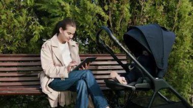 Trençkotlu bir kadın bankta bir tabletle meşgul, bebek arabası yakınlarda, açık havada huzurlu bir alışveriş anını ön plana çıkarıyor, kameralar sağa doğru dönüyor.