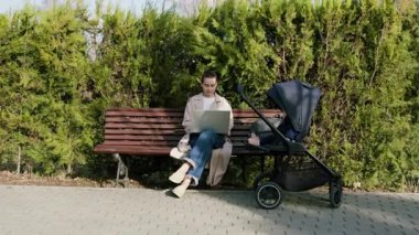 Kadın serbest çalışan, bilgisayarını bir park bankında, bebek arabasının yanında, yeşilliklerle çevrili olarak buluyor. Modern bir iş hayatı dengesini gösteriyor.