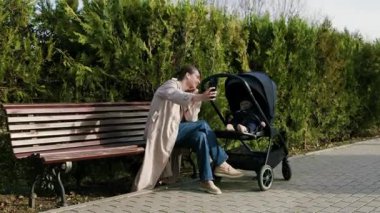 Bej ceketli bir kadın, park yolunda bebek arabasının yanında otururken akıllı telefonuyla neşeli selfie çekiyor, neşeli anı ve çevresini saran yemyeşil manzarayı vurguluyor