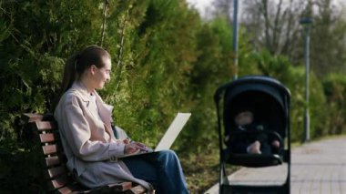 Açık renk ceketli bir kadın, yanında bir bebek arabasıyla bir bankta dizüstü bilgisayarıyla, dışarıda huzurlu bir çalışma ortamını resmediyor.