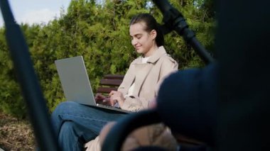 Bej renkli trençkotlu bir kadın, dizüstü bilgisayarıyla bir park bankında meşgul, yemyeşil, yoğun konsantrasyon ve sükunet gösteren kamerayla çevrili,