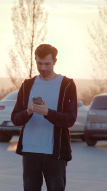 Erkek bireyler günbatımında park alanında akıllı telefonla etkileşime girerken, arka planda park edilmiş araçlar çağdaş araba paylaşım deneyimini göstermektedir.