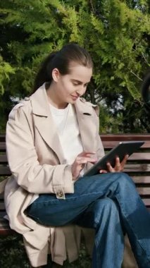 Trençkotlu bir kadın bankta bir tabletle meşgul, bebek arabası yakınlarda, açık havada huzurlu bir alışveriş anını ön plana çıkarıyor, kameralar sağa doğru dönüyor.