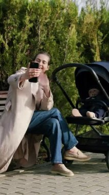 Kadın figürü, akıllı telefonuyla neşeli bir anı yakalıyor, bebek arabasının yanında oturuyor, yeşillikle çevrili, sıcaklığı ve bağlantıyı aktarıyor, kameralar yakınlaşıyor.