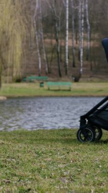 Bej ceketli kadın, ağaçlar ve yumuşak çimlerle çevrili, huzurlu bir göl kenarında bebek arabasıyla yürüyor. Sakin bir atmosfer ve doğal güzelliği ön plana çıkarıyor.