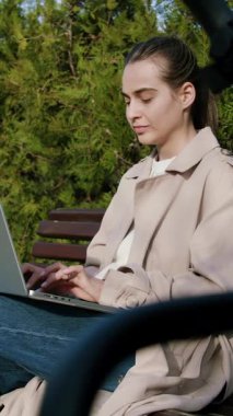 Trençkot giymiş bir kadın, yemyeşil yemyeşil bir bankta otururken aktif olarak dizüstü bilgisayardan online alışveriş yapıyor ve sakin bir doğa deneyimi sergiliyor.