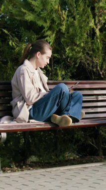 Kadın alışverişçi, yeşil alanlarla çevrili bir bebek arabasının yanındaki bankta otururken online alışveriş için akıllı telefon kullanıyor ve sakin bir alışveriş ortamı yaratıyor.