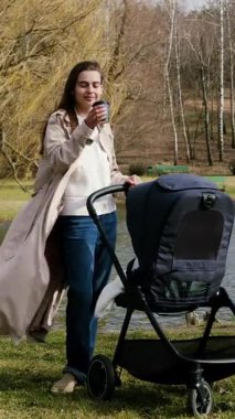 Paltolu kadın figürü, sakin bir göletin kenarında duruyor, bebek arabasını itiyor ve kahveyi yudumluyor, dışarıda huzurlu bir anı yakalıyor, kameralar sorunsuz bir şekilde hareket ediyor.