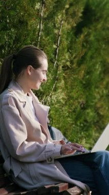 Açık renk ceketli bir kadın, yanında bir bebek arabasıyla bir bankta dizüstü bilgisayarıyla, dışarıda huzurlu bir çalışma ortamını resmediyor.