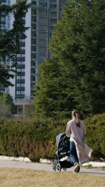Kadın figürü, yeşil ve kentsel yapılarla çevrili park ortamında bebek arabasıyla yürüyor, dingin atmosferi yakalıyor, kamera onu yol boyunca takip ediyor.