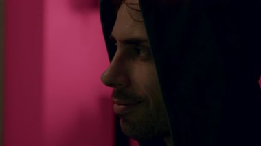 Koyu renk kapüşonlu bir erkek figürü, canlı pembe bir arka plana karşı ifadeler sergiliyor, duygusal derinliği ve karakter gelişimini vurguluyor.