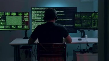 Erkek yapay zeka mühendisi, karmaşık verileri gösteren birçok ekranı olan bir masada çalışmaya dalmış, onun odaklanmasını ve etrafındaki teknolojik çevreyi vurguluyor.