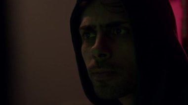 Karanlık teknoloji laboratuvarında kapüşonlu giyen Afrikalı Amerikalı bir adam kamera yaklaştıkça yoğun bir yüz ifadesi sergiliyor.