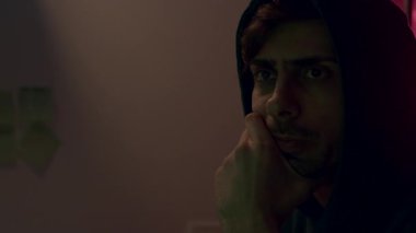 Kapüşonlu genç adam siber güvenlik konularına yoğun bir şekilde odaklanıyor. Loş bir ortamda, gölgeler atmosferi güçlendiriyor, kamera tavaları ve yakınlaştırıyor.