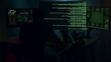 Dijital adli tıp uzmanı karanlık bir ortamda birden fazla monitör üzerinde veri analizi yapıyor, teknolojiyi ve karmaşık detayları vurguluyor, çalışma alanındaki kamera kalıpları