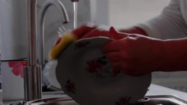 Kadın elleri aktif olarak mutfak lavabosundaki akan suyun altındaki tabağı yıkıyor, bulaşık yıkama işlemini ve su sıçramalarını vurguluyor, kamera detay için yakınlaştırıyor.