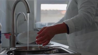 Beyaz elbiseli kadınlar mutfak lavabosunda bulaşık yıkamakla meşgul, akıcı su ve yansımalarla, temizlik ve ambiyansı vurguluyorlar.