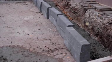 Devamlı inşaat sahnesinde, bir hendek boyunca yerleştirilen gri beton bloklar, kurulum sürecini ve çevreyi çevreleyen kameralı doli hareketler yer alıyor.
