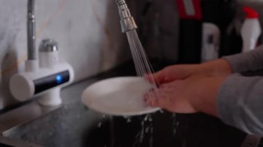 Kadın elleri aktif olarak akarsu altındaki bir tabağı yıkıyor. Pürüzsüz bir mutfak lavabosunda. Harekete yakınlaşan dinamik bir kamerayla temizlik sürecini vurguluyor.