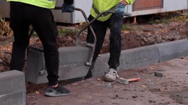 Yansıtıcı yelek giyen iki inşaat işçisi, yol boyunca beton kaldırma ve yerleştirme konusunda işbirliği yaparak, becerikli takım çalışması ve ekipman işleme becerilerini ortaya koyuyor