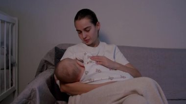 Bir anne bebeğini sevgiyle rahat bir kanepede emzirir. Duygusal bağlantıyı geliştirmek için yumuşak bir zum ile besleyici ilişkiyi ve sıcaklığı ön plana çıkarır.